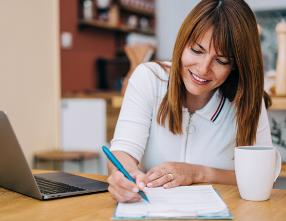 Image: Woman at table filling out a paper form with laptop open in front of her
